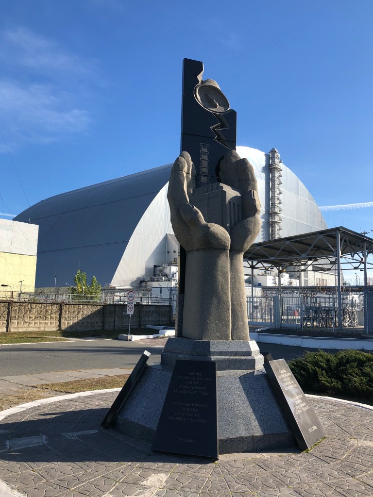 A statue of hands, holding a small power plant. Behind is the new confinement over the Chernobyl Power Plant.