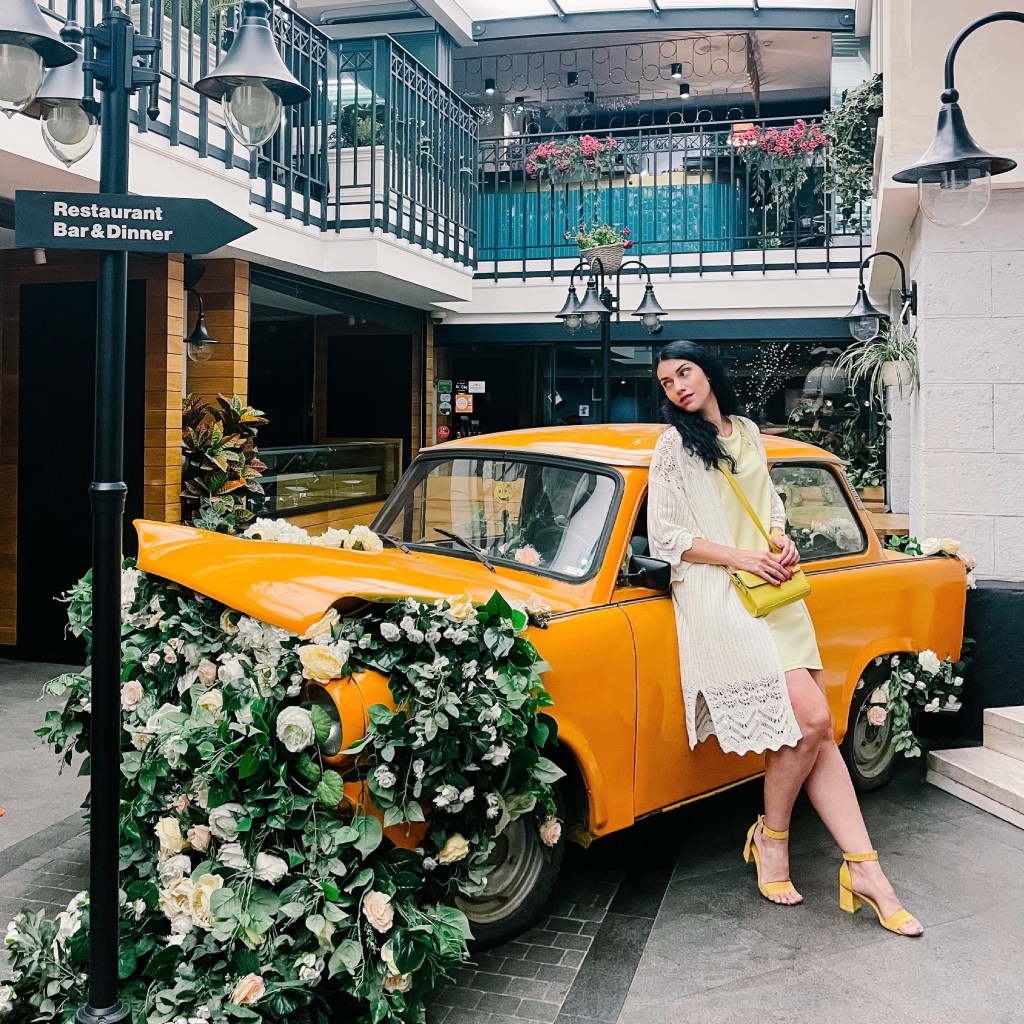 A picture of a brunette girl, standing next to an yellow car, full of flowers.